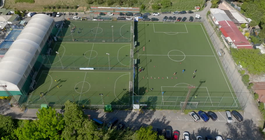 Aerial view of a vibrant sports complex featuring multiple artificial turf soccer pitches. The facility includes several smaller fields and a larger main pitch, a covered structure, and parking areas.