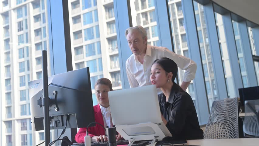 Colleagues of different ages and genders work together at desks with monitors and laptops, discussing projects against a backdrop of urban architecture