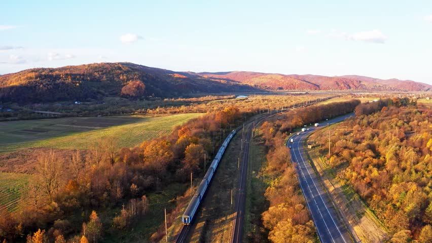 The train travels by rail to the distance of Carpathian mountains near the highway with cars in the forest in Ukraine