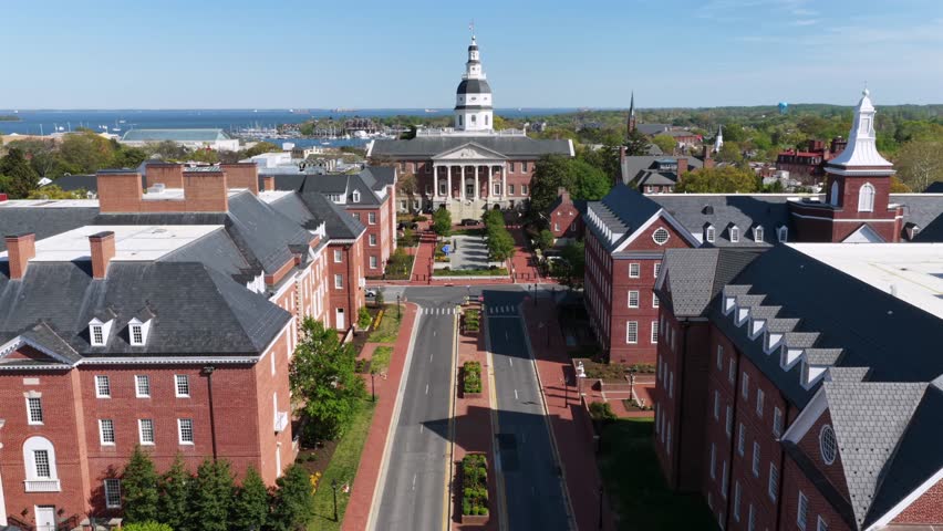 Aerial approach to Maryland State House in Annapolis, Maryland framed by red brick buildings and downtown streets, with Chesapeake Bay beyond under bright spring sunshine in historic USA capital city.