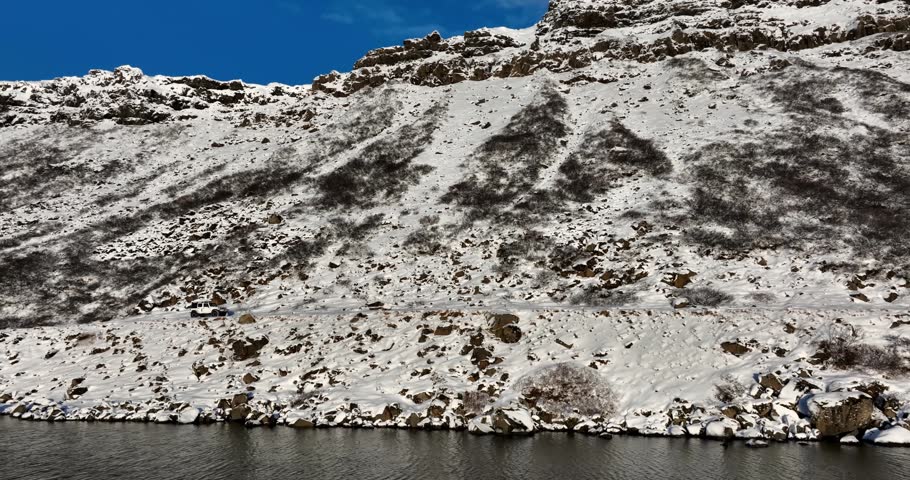 Aerial side view of a white 4x4 moving along the snowy cliffside shore of Kleifarvatn in Iceland, with rocky slopes and dark winter water below.