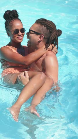 Vertical video: Laughing couple reacting to sun, hugging and drifting in home pool, orange swimwear. Water, affection, outdoor, sunlight, splash, sunglasses, vertical video