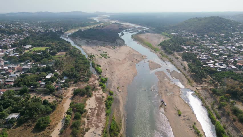An expansive aerial drone view showcases the partially dry river flowing through Tecpan de Galeana, Guerrero, revealing arid landscape and the surrounding residential town.