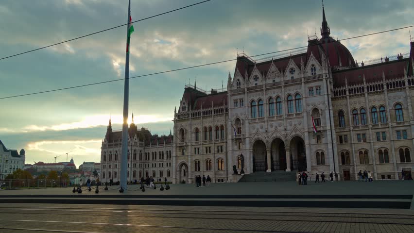 Yellow tram moving past the Hungarian Parliament Building in Budapest during a golden sunset with a tall flagpole and cloudy sky