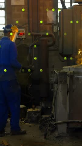 Vertical video: Worker stepping to forge finishing pour as sparks flying binary overlay hiding view. Foundry, metalworking, furnace, machinery, controls, workshop, vertical video