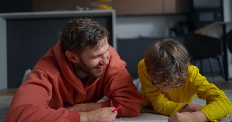 Smiling father and young child lying on a cozy living room floor drawing together, sharing playful learning time and warm family bonding at home
