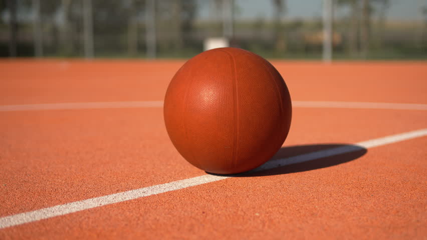Basketball ball lying on white line on basketball court in sunlight, gimbal shot. Arc shot of brown ball on sunny day on outdoor playing field without people.