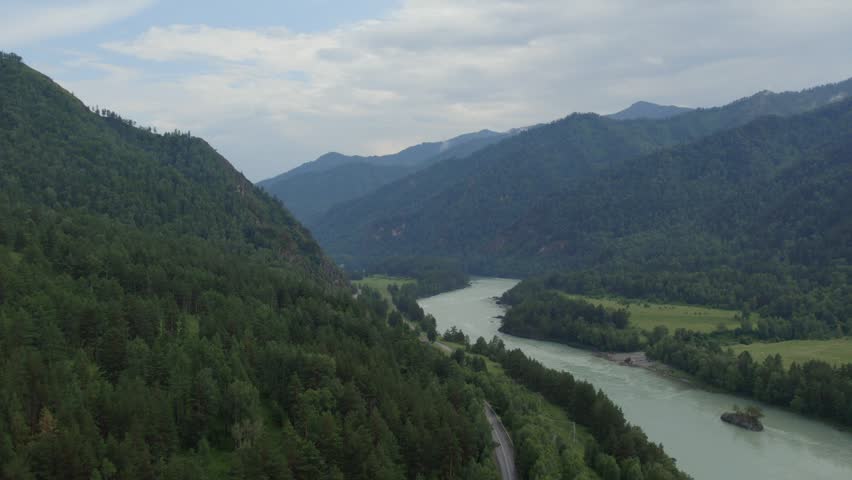 Drone footage of a winding turquoise river flowing through a green mountain valley and taiga forest in Altai, Russia, under a cloudy summer sky