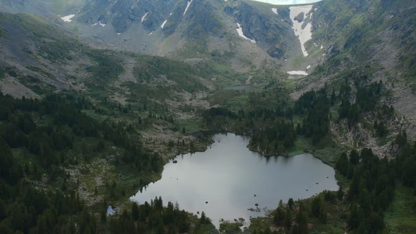 Drone footage of Karakol Lakes in the Altai Republic, Russia, surrounded by rocky peaks, alpine meadows, conifer forest and lingering snow patches in summer