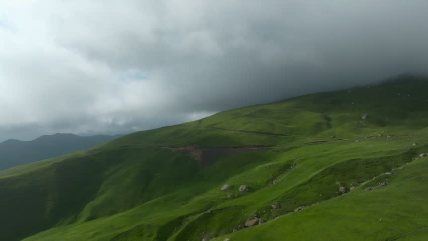 Wide landscape footage of lush green mountain slopes with a winding dirt road and dramatic low clouds after rain, filmed outdoors in Dagestan, Russia, Caucasus region