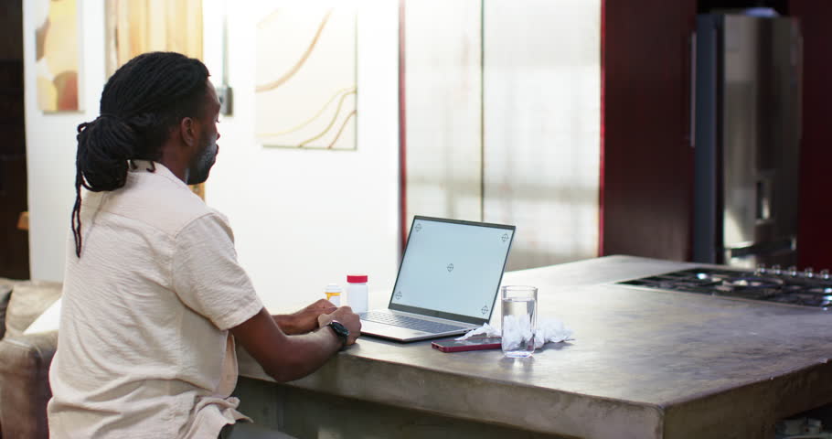 Man reacting to laptop, reaching for pill bottle with red cap, checking dosage at kitchen island. Person, home, countertop, reading, medication, container, focus