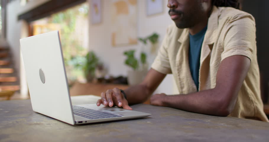 Man in overshirt tapping, typing on laptop at home, camera pulling back, reacting to message. Remote, workspace, cozy, warm, plants, bookshelf, art