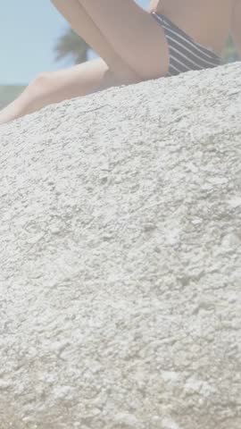Vertical video: Girl sitting rock, camera pulling back starting blue nodes causing leaning for tech. Child, beach, swimsuit, stone, outdoor, sky, vertical video
