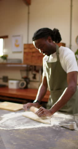 Vertical video: African American man in apron laying rolling pin, rolling dough for baking at bench. Baker, pastry, kitchen, wooden, flour, sunlight, vertical video