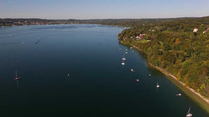Drone panorama of Starnberger See in Bavaria, Germany, golden autumn foliage, blue sky, bright sunlight, calm waves, yachts and sailing boats near the lakeshore, peaceful scenery.