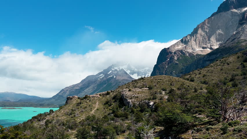 Rugged hiking trail overlooking the turquoise waters of Lago Nordenskjöld and towering granite peaks of Cuernos del Paine under fast-moving clouds in Torres del Paine, Patagonia, Chile.