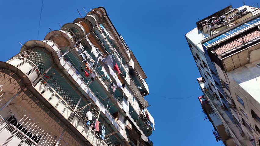 Low-angle view of weathered residential buildings with laundry hanging from balconies, revealing a traffic light against a clear blue sky in a crowded, rundown urban neighborhood