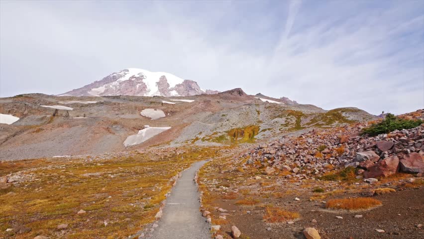 A scenic view of the trail leading to Panorama Point, with the snow-capped peak of Mount Rainier dominating the background. The alpine meadows display vibrant orange and yellow fall colors under a par