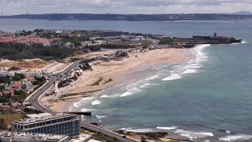Aerial view of Carcavelos beach with waves crashing on the shore, coastal road, and Sao Juliao da Barra Fort in Lisbon, Portugal.
