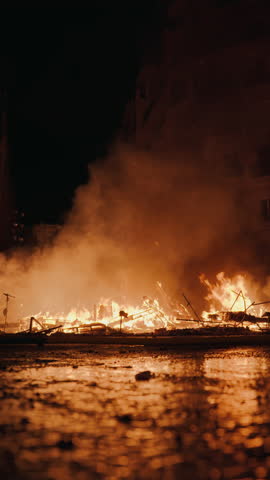 Ground level view of burning Las Fallas falla debris during La Crema in Valencia, flames and glowing embers reflected in wet cobblestone pavement with dramatic smoke rising into dark sky