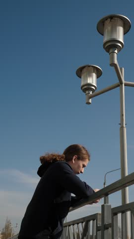 Young person leaning on railing by city lamppost, student checking phone, curly hair in hooded jacket, metal railing and clear blue sky, midday light with soft shadows, quiet urban moment of solitude