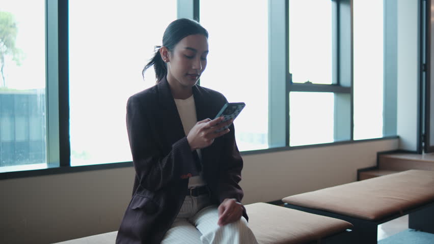 One young, attractive Asian businesswoman in suit, online chatting and browsing on a mobile phone at modern workplace lounge, financial job, and professional communication, urban technology lifestyle.