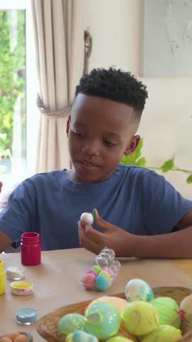 Vertical video: Picking up foam eggs, boy in blue tee tasting painted piece decorating eggs at home. Kid, craft, pastel, art, family, window, vertical video