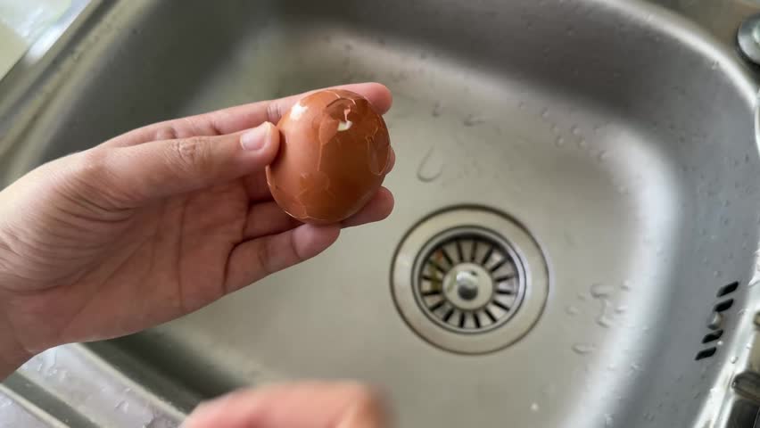 Hands peeling a boiled egg over a kitchen sink, showing a simple step in food preparation.