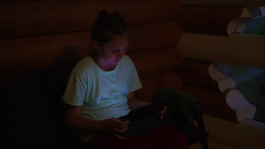 Young girl sits in a dark log cabin, focused on her tablet. She wears a white shirt and red pants, hunched over her device. Warm wooden walls surround her, lit by soft ambient light