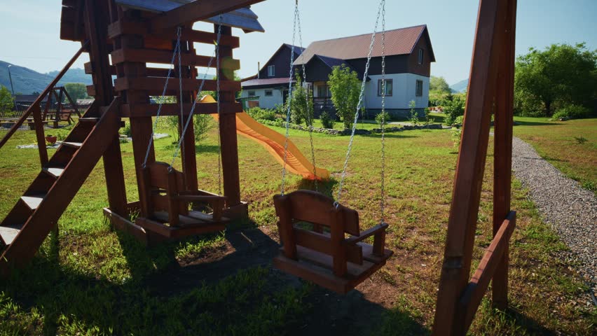 Wooden swing set stands in a grassy yard. Swings hang still under bright sunlight. House with a sloped roof appears in background. Gravel path leads through green lawn. Scene feels calm and peaceful