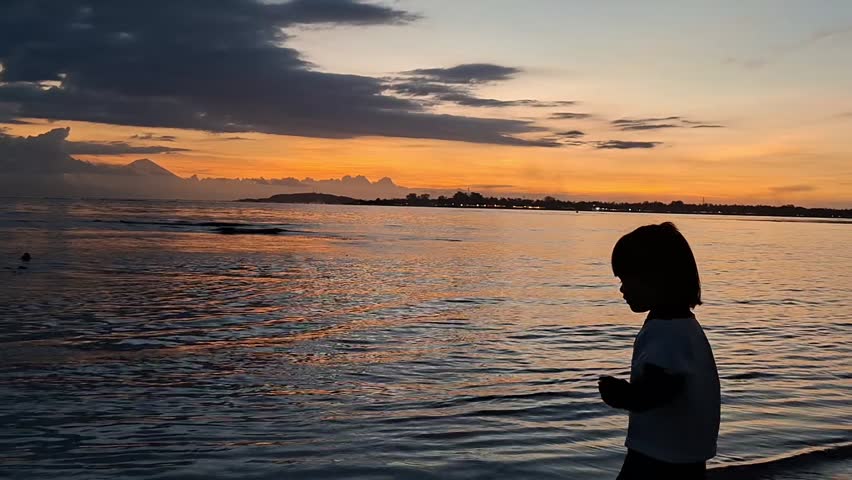 Silhouette of a young child standing on a sandy beach at sunset and throwing a stone into the calm ocean water with a vibrant orange sky in the background.