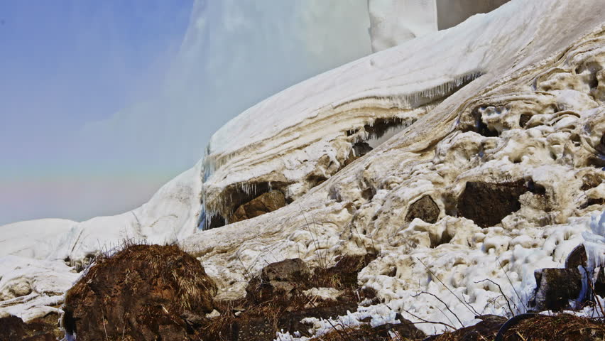 Snow covers rocks at the edge of a river, with ice hanging from the surface. The sky is clear, and the scene shows winter in a forested region.