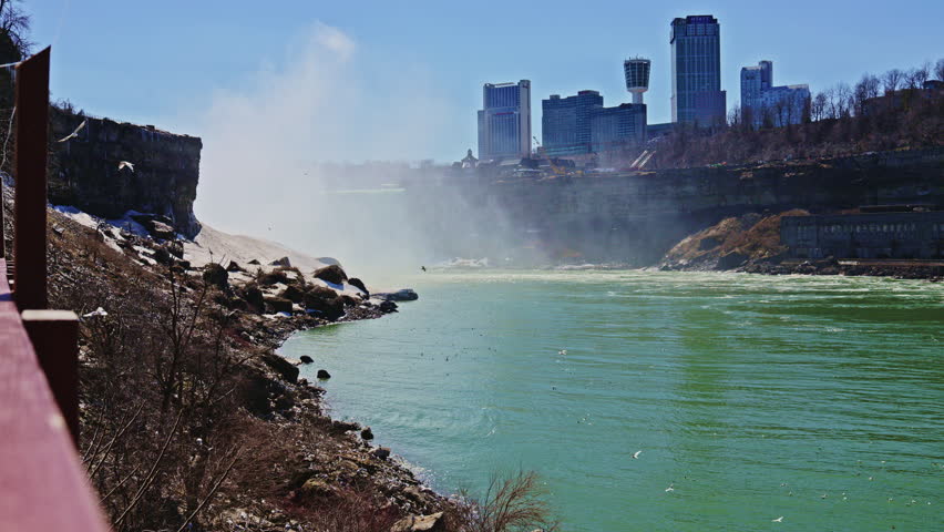 People stand near the water as the river flows fast towards the falls. Mist rises from the water while buildings appear in the distance against a blue sky.