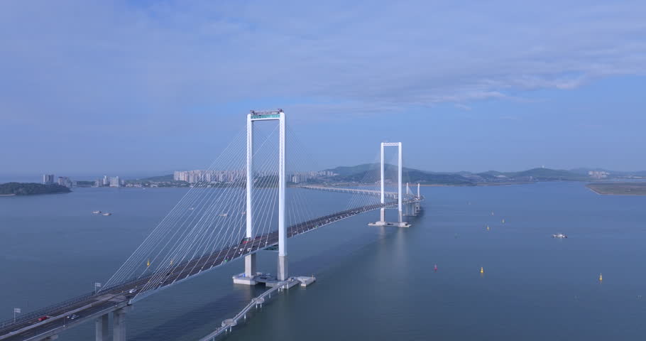 Cinematic aerial view of Cheongna Sky Bridge in Incheon, South Korea. The large cable-stayed bridge stretches across the blue sea, creating a modern coastal landscape with transportation infrastructure and ocean scenery.
