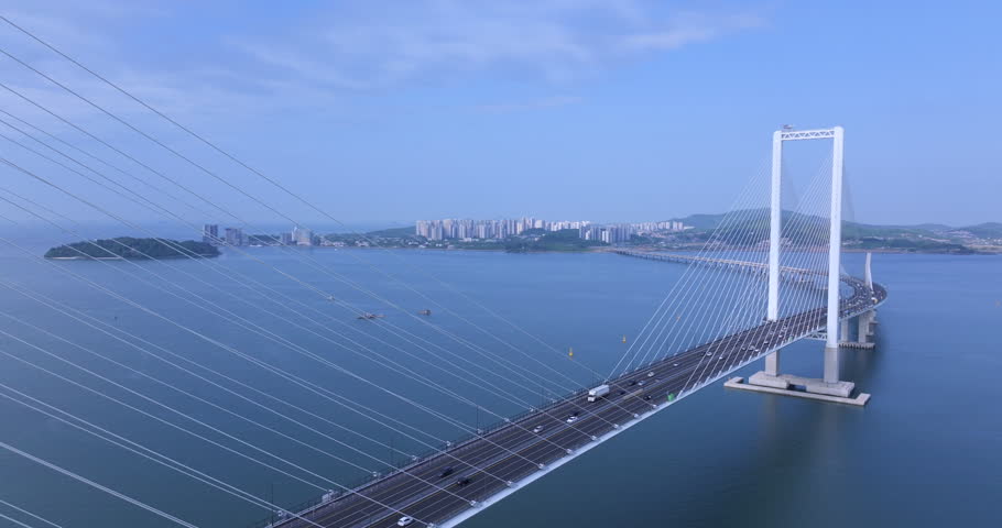 Cinematic aerial view of Cheongna Sky Bridge in Incheon, South Korea. The large cable-stayed bridge stretches across the blue sea, creating a modern coastal landscape with transportation infrastructure and ocean scenery.
