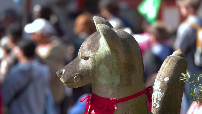 Close-up of a stone fox statue with red bib before a blurred crowd of tourists