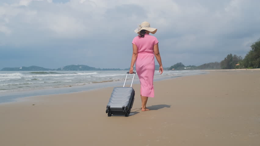 Young traveler in straw hat walking along sandy beach and pulling rolling suitcase near sea waves during tropical vacation, slow motion. Holiday travel concept