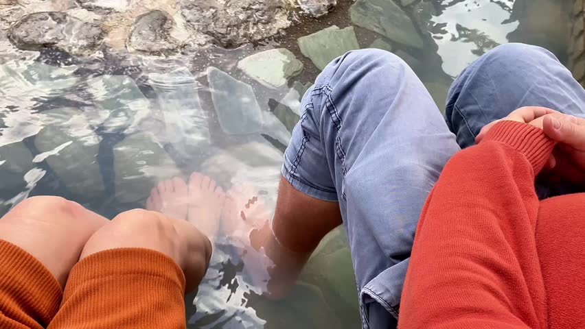 A high-angle shot showing the legs and feet of a man and a woman as they soak in a natural stone-lined hot spring. The clear, warm water flows over smooth grey rocks, with gentle steam rising from the surface. This candid moment captures shared relaxation, wellness, and tranquility in a traditional thermal spa setting