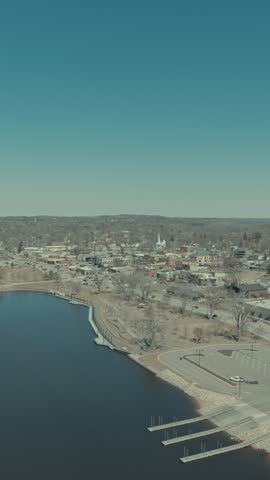 Aerial view from over calm St Croix River toward downtown Hudson Wisconsin showing curved shoreline, waterfront promenade, marina docks, church steeple and bare spring trees