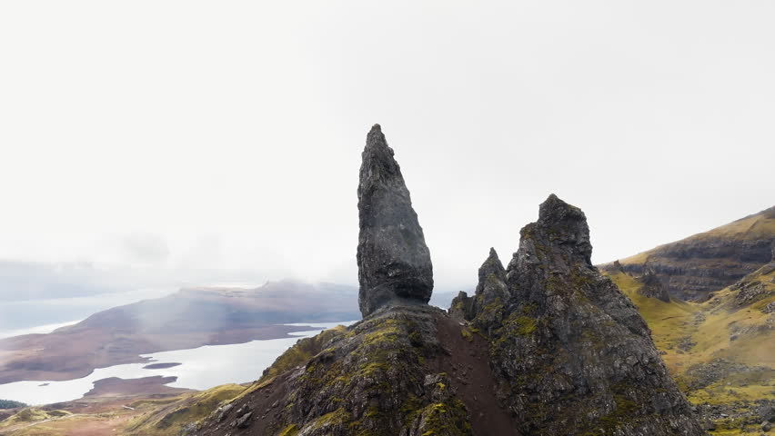 Sweeping drone shot circling the Old Man of Storr, Scotland, cloud and mist rendering the landscape monochromatic and hauntingly beautiful, dramatic natural light conditions throughout