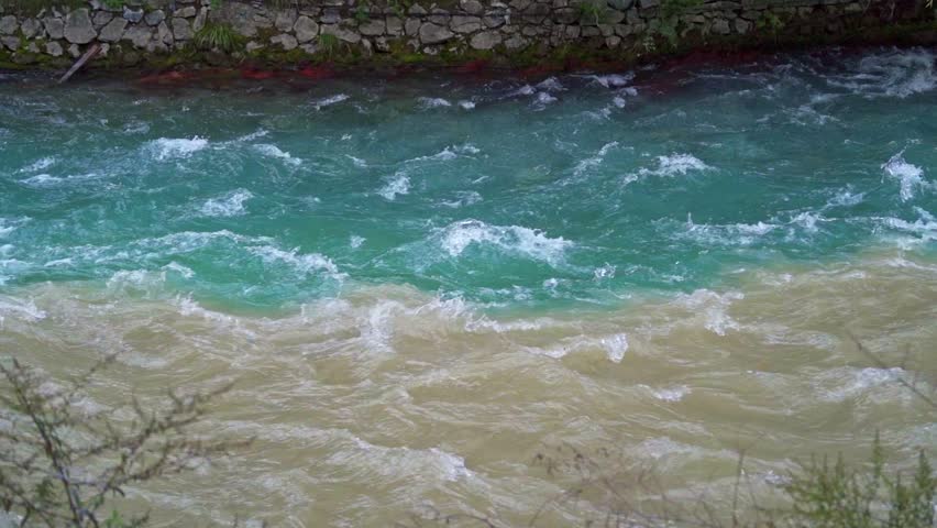 Two mountain streams merging in Jiuzhaigou National Park. The shot captures the turbulent mixing zone between clear turquoise water and murky brown sediment-filled runoff against a stone embankment.