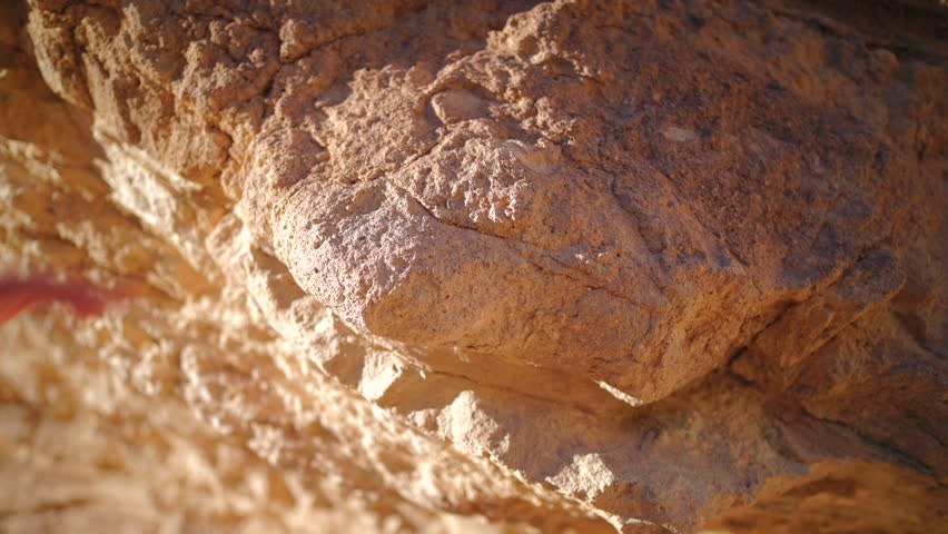 Close-up of a hand gently touching and feeling the textured surface of a rugged sandstone boulder bathed in golden sunlight. Warm orange and tan tones highlight the weathered rock formation.