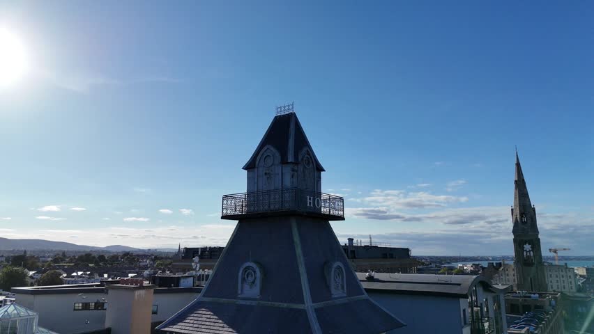 Cinematic aerial orbit shot of the distinctive rooftop Hotel in Dún Laoghaire, Dublin, Ireland. Bright sunny day with clear blue skies overlooking Dublin Bay and the Irish Sea. Elegant coastal architecture and seaside town atmosphere captured in smooth drone motion.
