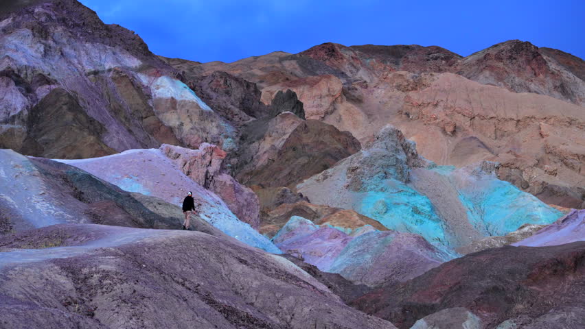 Lone hiker stands amid vibrant multicolored volcanic rock formations at Artists Palette in Death Valley National Park. Purple, turquoise, orange and pink mineral deposits create surreal landscape.