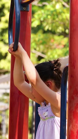 A young girl hangs from blue rings at a sunny outdoor playground in Lumpini Park