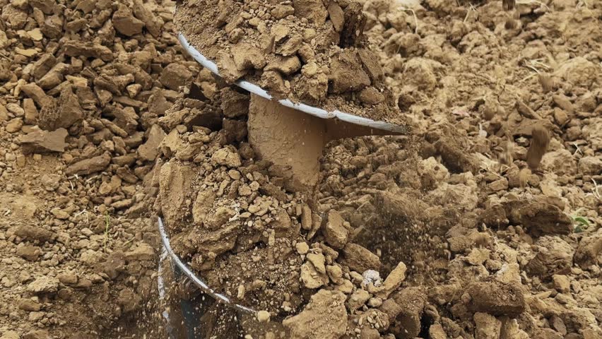 Close view of auger drill bit coming out of ground after completing hole, soil and debris falling off with tool visible in a rural outdoor construction setting