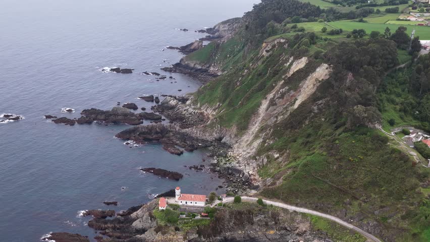 Aerial view of the Faro de Cudillero lighthouse perched on a rugged cliffside overlooking the Cantabrian Sea with lush green hills in Cudillero, Asturias, Spain.