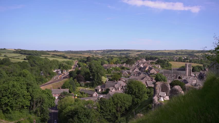 Panoramic View of Historic Corfe Castle Village and Dorset Countryside

A cinematic panoramic shot of the historic village of Corfe Castle in Dorset, England, nestled at the foot of ancient ruins under a clear blue summer sky, showcasing the traditional stone architecture and the lush, rolling green landscape of the Purbeck countryside.