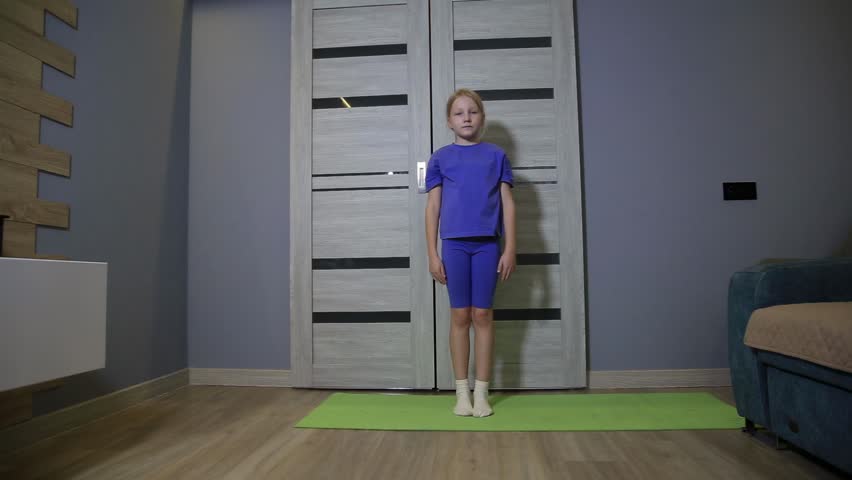 Full shot of young caucasian girl in blue sportswear performs warm-up exercises on green yoga mat at home. Child raises arms, lifts leg, focusing on body movement, health.