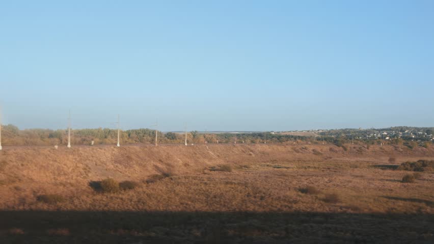 Scenic view from moving train, revealing extensive landscape of railway track, electric poles, and autumnal foliage. Dry golden grass covers side of embankment, leading to distant small town.
